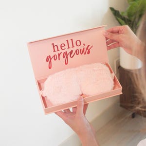 Może przedstawiać: R&oacute;żowe pudełko z nadrukiem "hello, gorgeous" na pokrywce. Pudełko jest otwarte i zawiera r&oacute;żową, puszystą maskę na oczy.