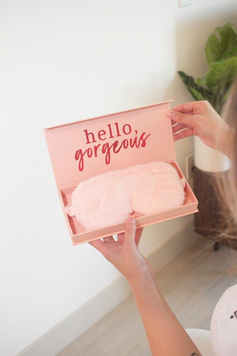 Może przedstawiać: R&oacute;żowe pudełko z nadrukiem "hello, gorgeous" na pokrywce. Pudełko jest otwarte i zawiera r&oacute;żową, puszystą maskę na oczy.