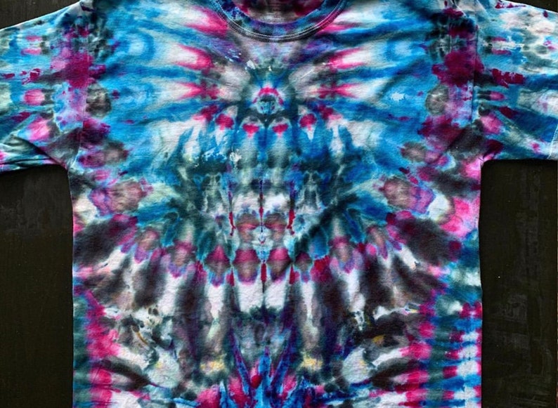 Puede incluir: Una camiseta tie-dye con un esquema de color azul, rosa y negro. La camiseta tiene un dise&ntilde;o sim&eacute;trico con un punto central y l&iacute;neas radiantes.