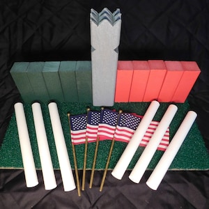 May include: A set of miniature American flags on wooden sticks, with white poles and green grass in the background. There are also several green, red, and blue wooden blocks in the background.
