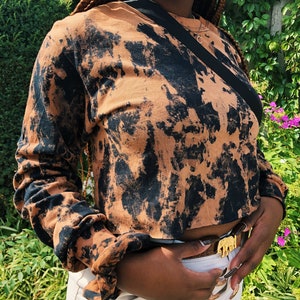 May include: A brown and black tie-dye long-sleeved shirt with a cropped hem. The shirt is worn with a white skirt and a gold belt featuring an elephant design.