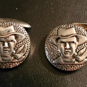 May include: A pair of silver cufflinks featuring a detailed embossed image of a man wearing a hat. The cufflinks have a textured circular design.
