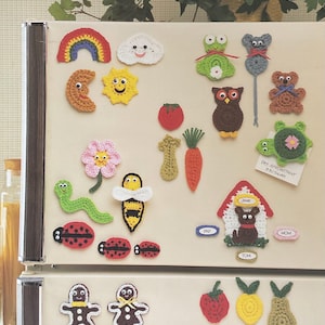 May include: A selection of handmade crochet items, including a rainbow, sun, and cloud, alongside animal and food designs. The image features a frog, owl, bear, tomato, and carrot. The text "25 DESIGNS TO CROCHET" is visible at the top.