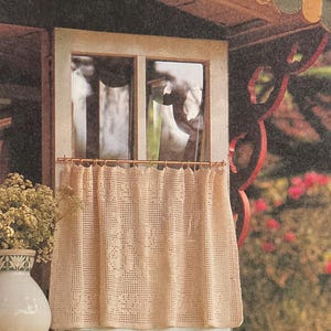 May include: A cream-colored, crocheted curtain hangs in front of a window. The curtain is patterned with openwork designs. A vase with dried flowers sits on the windowsill. The window is part of a small, decorative structure with a green and yellow base.