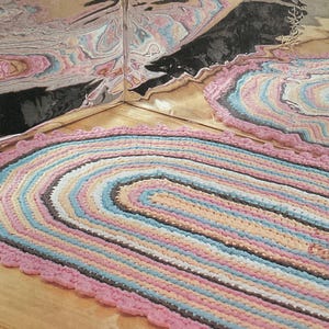 Può includere: Tappeto ovale intrecciato con bordo smerlato. Il tappeto presenta una palette di colori pastello rosa, blu, pesca e marrone. Il tappeto è posizionato su una superficie di legno e un angolo di un muro a specchio è visibile sullo sfondo.