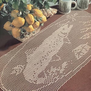 May include: A cream-colored, crocheted table runner with a central floral design. The runner is placed on a brown surface, with a basket of lemons and two mugs with ship illustrations in the background.