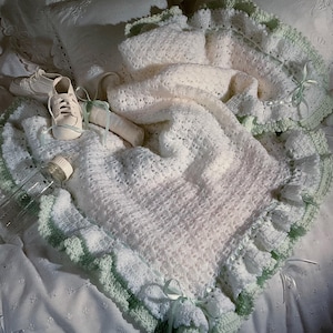 Puede incluir: Una manta de bebé blanca hecha a mano, tejida a ganchillo, con un ribete verde claro y detalles de lazo. La manta está sobre una colcha blanca, con zapatos de bebé blancos, un biberón y una manta enrollada.
