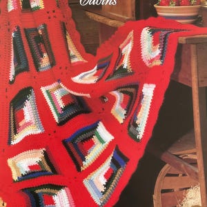 Könnte beinhalten: Eine leuchtend rote gehäkelte Decke mit farbenfrohen quadratischen Hüttendesigns. Die Decke hat einen gewellten Rand und ist über eine Holzoberfläche drapiert. Der Text "Colorful Cabins" ist sichtbar.