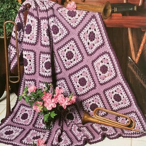 May include: A handmade crochet blanket in shades of purple and white, featuring a square pattern design. The blanket is draped over a wooden surface, with a trumpet and flowers adding decorative elements. The blanket's dimensions are not visible.