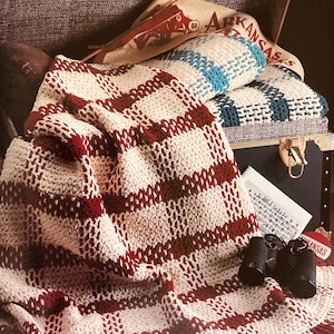 May include: A red and cream checked woven blanket with a fringed edge is draped over a trunk. Inside the trunk are striped pillows in blue and white, and a pillow with a pig graphic.