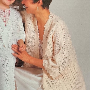May include: A mother and child are wearing matching cream-colored, open-front crochet cardigans. The cardigans have a scalloped edge and a delicate, open weave. The child is smiling. The mother is smiling and has short, dark hair.