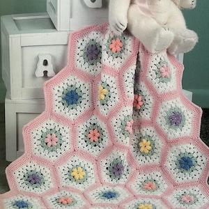 Puede incluir: Una manta de bebé hecha a mano con ganchillo, con un patrón hexagonal y un borde rosa. La manta está adornada con aplicaciones de flores coloridas en tonos de azul, amarillo, rosa y morado. Un oso de peluche blanco con un lazo rosa está encima.