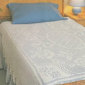 May include: A white, intricately crocheted bedspread with a floral and geometric pattern covers a bed. A blue pillow rests on the bed, and a wicker headboard is visible. A blue lamp sits on a nightstand.