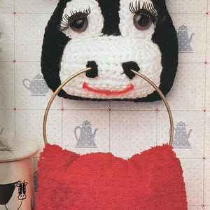 May include: A whimsical, handmade cow-shaped towel holder. The black and white crocheted cow face features large eyes with long eyelashes and a red smiling mouth. A gold-colored metal ring holds a bright red towel.