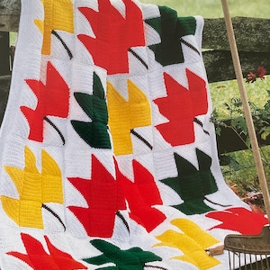 May include: A crocheted blanket featuring a leaf pattern in red, yellow, and green on a white background. The blanket is labeled "Fall Foliage" and is part of the "Afghan Collector's Series".