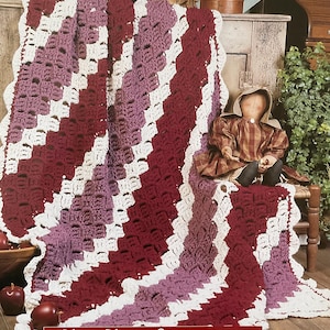 May include: A crochet blanket with a zig-zag design in white, burgundy, and lavender stripes. The blanket has a scalloped edge. The text "Zig-Zag Shells" is displayed at the bottom of the image.