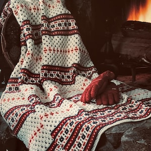 May include: A cream-colored crocheted blanket featuring horizontal bands of red and navy blue geometric patterns, draped over a surface. A pair of red insulated gloves rests on top of the blanket, and a red boot is visible in the lower corner.