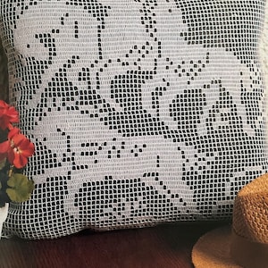 May include: A square pillow with a white crocheted design on a black background. The crocheted pattern appears to depict a floral or leaf motif. The pillow is resting on a dark wooden surface, with a straw hat visible in the background.