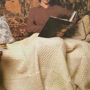 May include: A cream-colored, textured knitted blanket with various stitch patterns. The blanket is draped over a person reading a book. The background features a tapestry with figures and floral designs. Pillows are on either side.