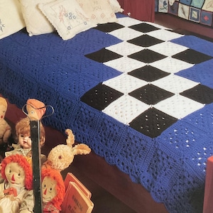 May include: A handmade blue, black, and white crochet blanket with a diamond pattern. The blanket is on a bed with decorative pillows. The blanket's edge has a scalloped design. Dolls and books are in the foreground.