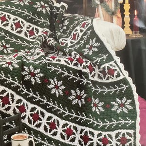 May include: A green and white crocheted afghan with red poinsettia flower designs. The afghan is draped over a sofa. A small chair holds a mug and a pair of burgundy slippers with bows are on the floor. The text "Poinsettia Afghan" is at the top.