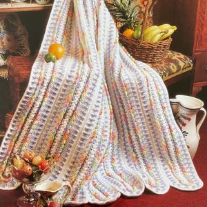 Peut inclure: Une couverture crochetée blanche et pastel avec un bord festonné. La couverture présente un motif texturé et est drapée sur une étagère avec des fruits et un panier. Le texte "Hawaiian Dreams" est en bas.
