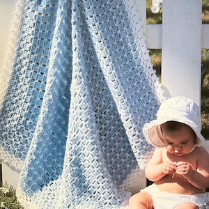 May include: A handmade, light blue and white crochet baby blanket with a textured pattern. The blanket is draped over a white picket fence. A baby wearing a white sun hat and diaper sits on the grass.