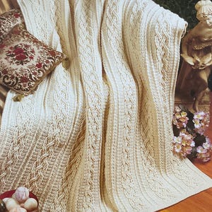 May include: Cream-coloured, intricately woven afghan blanket with a cable knit design. The blanket is draped over a sofa, with a decorative pillow featuring a floral pattern. The text "Interwoven Cables" is visible.