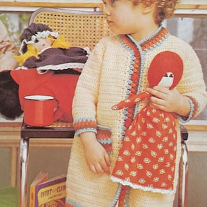 May include: A child wearing a cream-colored crochet cardigan with blue and orange trim, holding a red doll with a patterned dress. The doll has a red head and white face. A doll and a red mug sit on a chair.