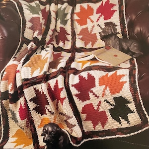 Op de afbeelding: Een gehaakte deken met een bladerenpatroon in herfstkleuren rood, oranje, geel en groen. De deken heeft een bruine en crèmekleurige rand, met de tekst "Autumn Leaves" onderaan. Hij ligt over een bruine leren bank.