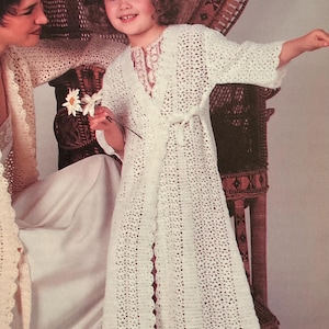 May include: A young girl wearing a white, open-front crochet robe with a tie belt. The robe has a scalloped hem and is paired with a lace-trimmed top. She holds white flowers. A woman is seated nearby, also wearing a crochet garment.