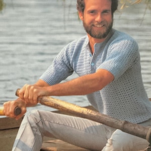 May include: A light blue knitted shirt with short sleeves and a button-up collar. The shirt has a textured pattern. The person is wearing light grey trousers and is in a wooden rowing boat on a lake. The person is holding oars.