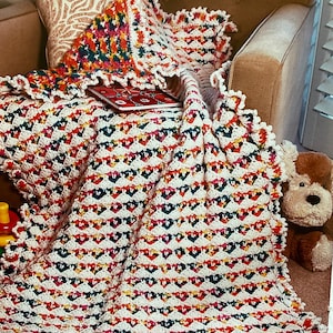 May include: A colorful, handmade crochet blanket with a scalloped edge. The blanket features a repeating pattern of white, red, yellow, green, and blue stitches. A matching pillow and a small stuffed dog are also visible.