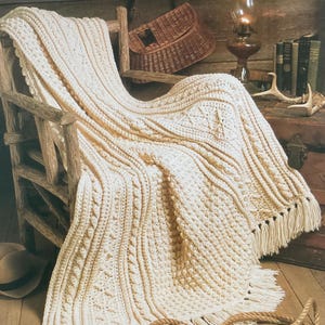 Może przedstawiać: Kremowy, ręcznie robiony koc z teksturowanym wzorem i frędzlami, narzucony na drewniane krzesło. Koc zawiera napis "Fisherman's Blanket" w eleganckiej czcionce. Scena przywołuje przytulną, rustykalną estetykę.