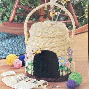 May include: A cream-coloured crocheted beehive yarn holder with a black entrance, adorned with flowers, ribbons, and bee embellishments. Features a handle and two bee-topped knitting needle holders. Yarn balls and a crochet tool organiser are also visible.