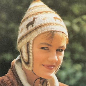 Puede incluir: Un gorro de punto con orejeras y una parte superior puntiaguda. El gorro presenta un patrón repetido de siluetas de perros salchicha en colores marrón y crema. El gorro se usa con una bufanda color crema y una chaqueta de pana marrón.