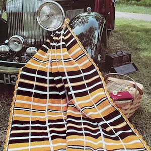 May include: A crocheted blanket with a plaid pattern in black, white, and yellow. The blanket is draped over the front of a vintage black car. A picnic basket and a small red book are in the background.