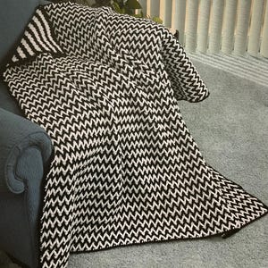 Könnte beinhalten: Eine schwarz-weiß gemusterte Decke mit Zickzackmuster, die über einem blauen Sessel drapiert ist. Das Design der Decke weist ein sich wiederholendes Zickzackmuster auf. Die Decke besteht aus einem weichen Material und eignet sich als Überwurf oder Dekoration.