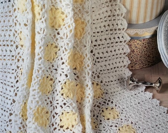 May include: A white crochet blanket with yellow flower motifs. The blanket features a scalloped border and is draped. The open weave and floral pattern give a delicate, vintage feel. A cosy home decor item, the blanket is likely handmade.