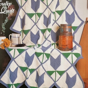 May include: A crocheted quilt with a tulip pattern in white, blue, and green. The quilt is folded over a surface, showcasing its geometric design and the text "Tulip Quilt" in the upper left corner. The quilt is part of the "Afghan Collector's Series".
