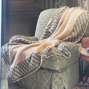 May include: A cozy armchair draped with a beige and green patterned throw blanket. The blanket features a textured design with a contrasting border. The chair is upholstered in a light green fabric with a subtle diamond pattern, creating a warm and inviting atmosphere.