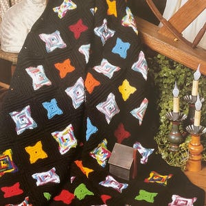 以下が含まれることがあります： カラフルな正方形の模様が入った、黒色の手編みのアフガンブランケット。正方形には、赤、黄、青、オレンジなど、さまざまな色の星のようなデザインが施されています。「Cathedral Window」の文字が下部に表示されています。