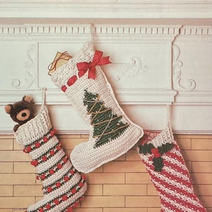 May include: Three crocheted Christmas stockings hanging on a fireplace. One has a teddy bear, another a Christmas tree, and the third red and white stripes. Festive, handmade decorations.