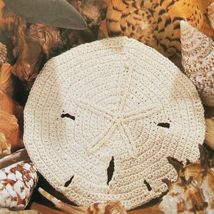 May include: Cream-colored, crocheted sand dollar with a star design in the center and several small holes. The sand dollar is surrounded by various seashells in shades of brown, tan, and white, creating a beachy theme.