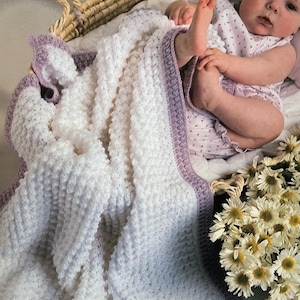 May include: A white and lavender crochet baby blanket with a textured pattern. The blanket is in a wicker basket with a baby wearing a light purple outfit. A bouquet of white daisies is next to the basket.