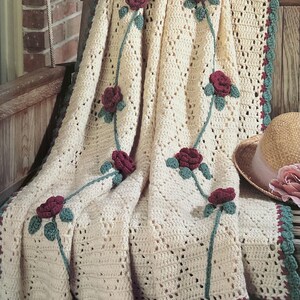 Puede incluir: Manta de ganchillo color crema con diseño de rosas. La manta presenta rosas rojas oscuras con hojas y tallos verdes. Los bordes están adornados con un borde festoneado en verde y burdeos.