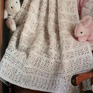 Puede incluir: Una manta de bebé blanca tejida a ganchillo con un patrón moteado de colores pastel. La manta está sobre una silla de madera, con un conejito y un cordero de peluche cerca. Los bordes de la manta tienen un borde decorativo.