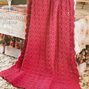 May include: A raspberry-coloured crocheted afghan blanket with a scalloped edge is draped over a bed. The blanket is made of a textured yarn and has a gradient effect, transitioning from a darker to a lighter shade of red. The text "Raspberry Creme" is visible.