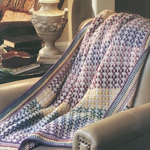 May include: A colourful crochet blanket draped over a leather armchair. The blanket has a pattern of white and coloured stitches in shades of purple, red, yellow, and blue. The edges are finished with a striped border.