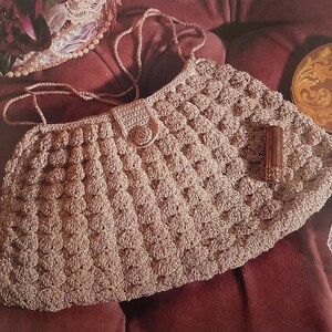 May include: A beige crocheted handbag with a textured, raised pattern. The bag has a flap closure with a button and a long strap. A tube of lipstick rests on the bag. The bag is on a maroon surface.